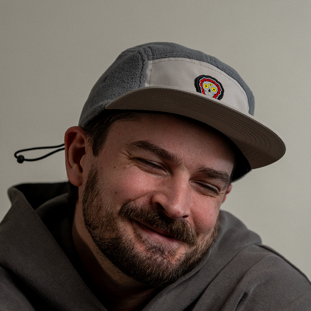 Front angled view of model smiling wearing Skull Flower Fleece 5 Panel hat showing PVC skull flower patch