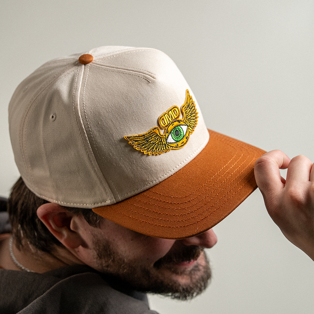 Model adjusting brim of Flying Eye Two-Toned Hat showing embroidered green eye patch