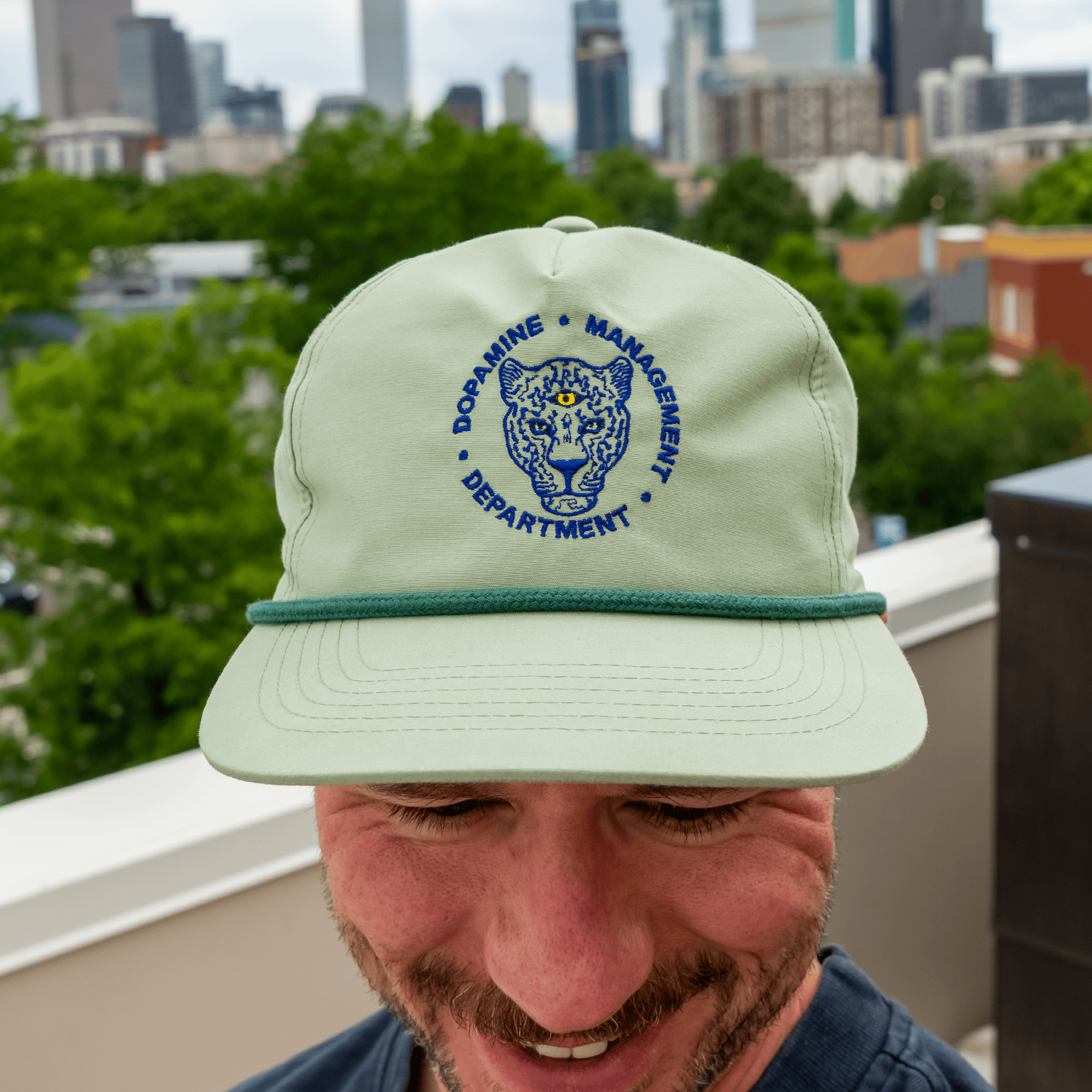 Person wearing a light green cap with a logo and text, outdoors with trees and buildings in the background