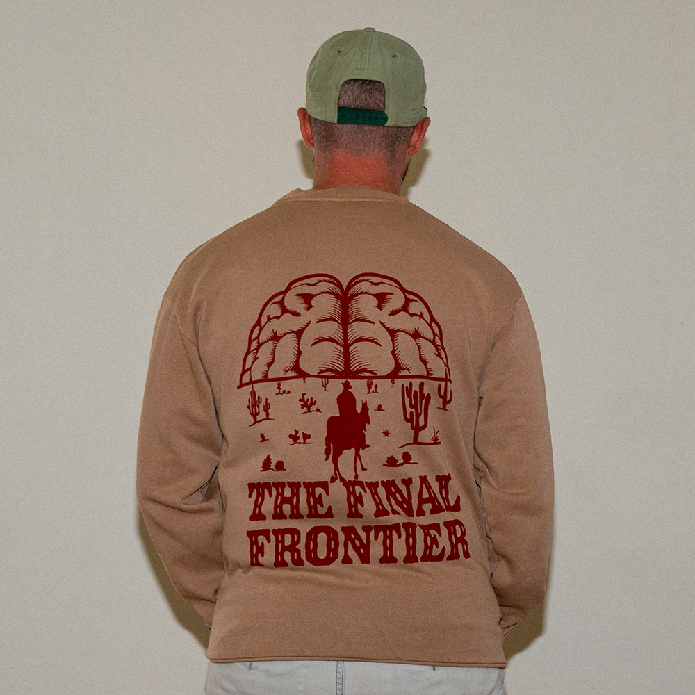 Model wearing tan Frontier Heavyweight Crewneck with oversized cowboy and brain mountain graphic on back