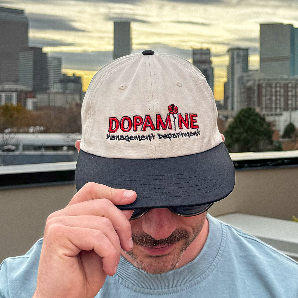Person wearing the two-tone cream and black Rose Logo Hat with embroidered Dopamine Management Department logo outdoors.