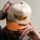 Model adjusting brim of Flying Eye Two-Toned Hat in sunlight, showing embroidered green eye patch