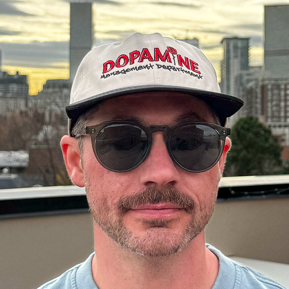 Close-up of the Rose Logo Hat worn by a model with sunglasses in front of a sunset skyline.