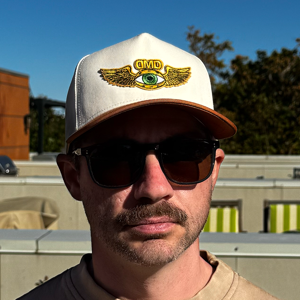 Close-up of model smiling in DMD Flying Eye Two-Toned Hat featuring gold winged eye embroidery