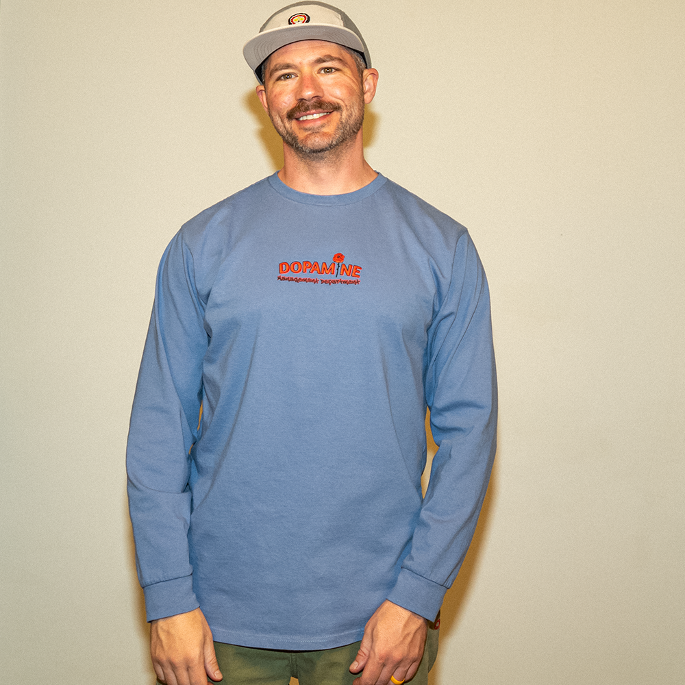 Model smiling in blue Kindness Long Sleeve Tee and grey hat – front logo view.