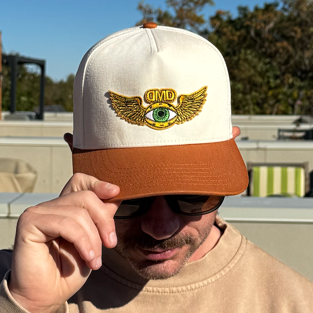 Model adjusting brim of Flying Eye Two-Toned Hat in sunlight, showing embroidered green eye patch