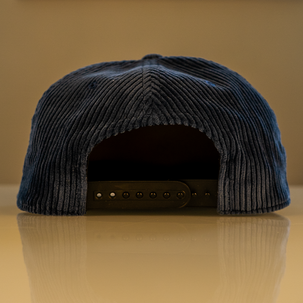 Navy blue corduroy cap with black snapback closure on a reflective surface with a beige background