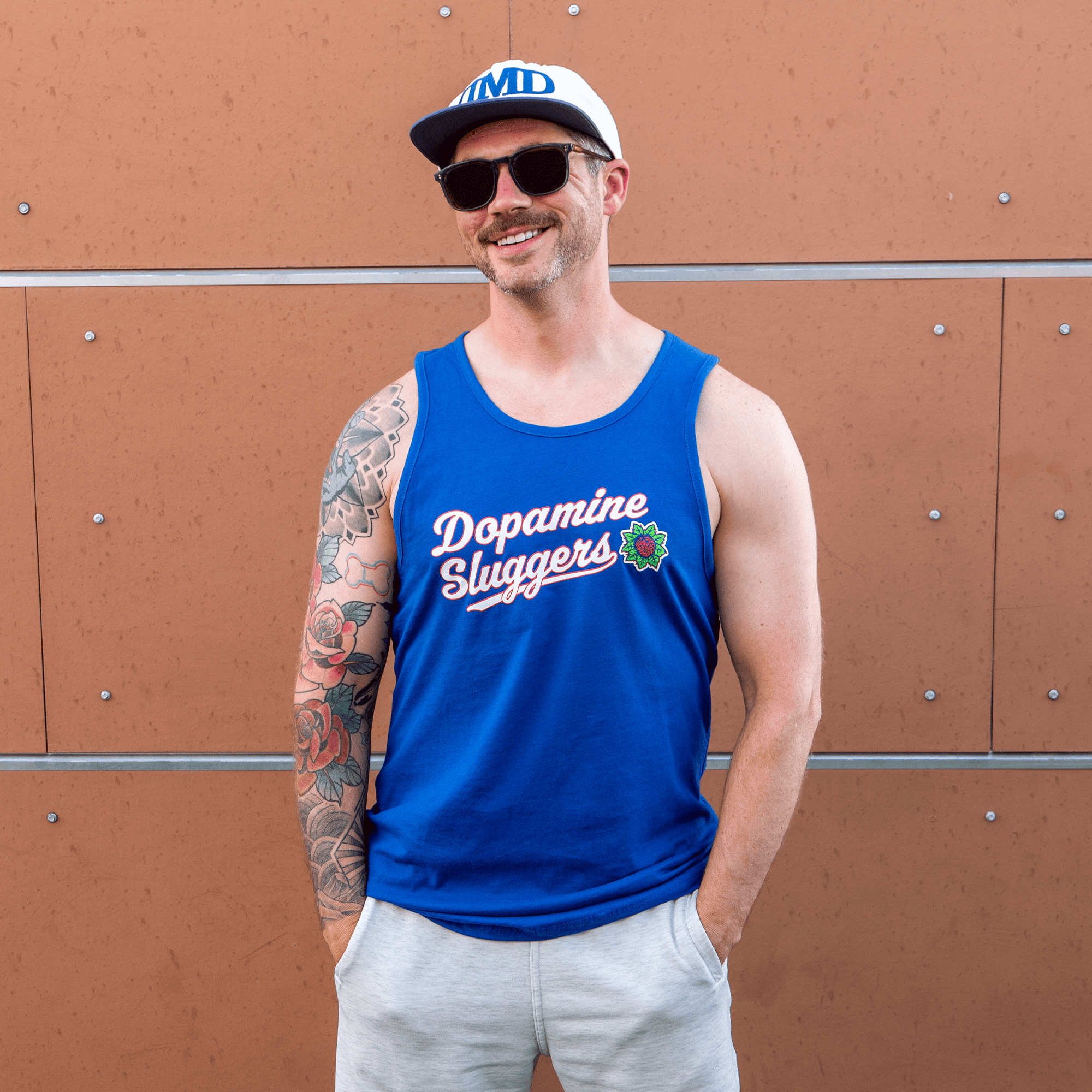 Tattooed man in Dopamine Sluggers tank top standing against modern orange wall