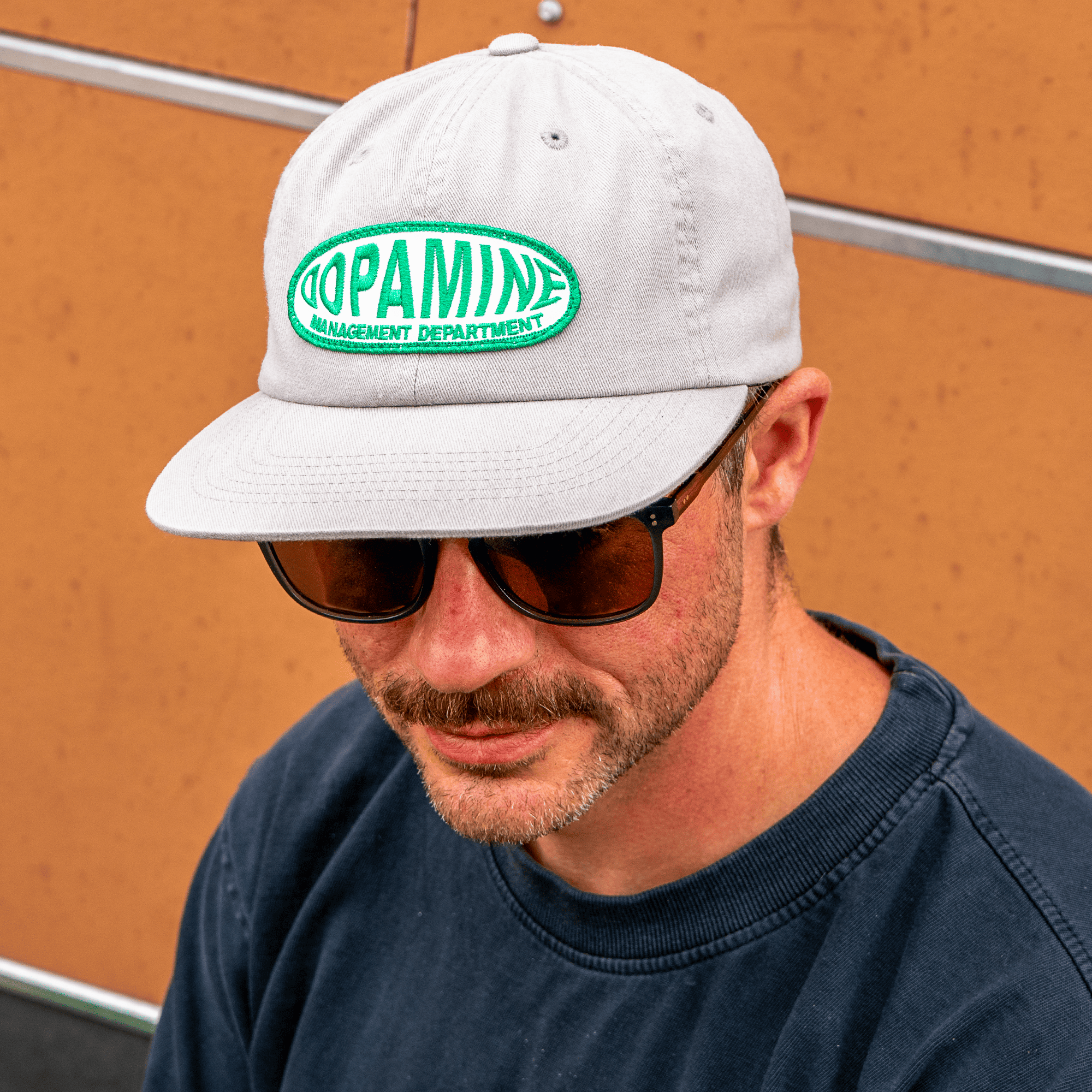 Man wearing Dopamine Management Department Warp Patch Hat and sunglasses, against an orange wall.